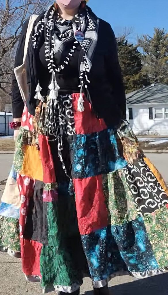 A picture of a person in a big patchwork skirt, a black cardigan sweater, and a keffiyeh with a tote bag under their arm.